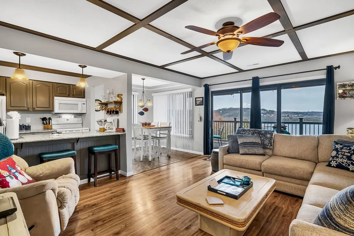 A spacious living area with comfortable seating, a kitchen with bar stools, and large windows providing a sweeping view of the lake. The light-filled room invites relaxation, family gatherings, and entertaining guests. The connection to the outdoors is strong, with the deck just beyond the windows for easy access to fresh air.