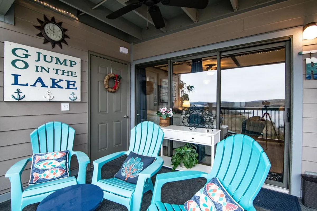 A welcoming outdoor patio with blue Adirondack chairs and a decorative sign, offering a peek through the glass doors and a glimpse of the natural surroundings. The setup invites relaxation and group gatherings, perfect for family downtime or morning coffees before a day out.