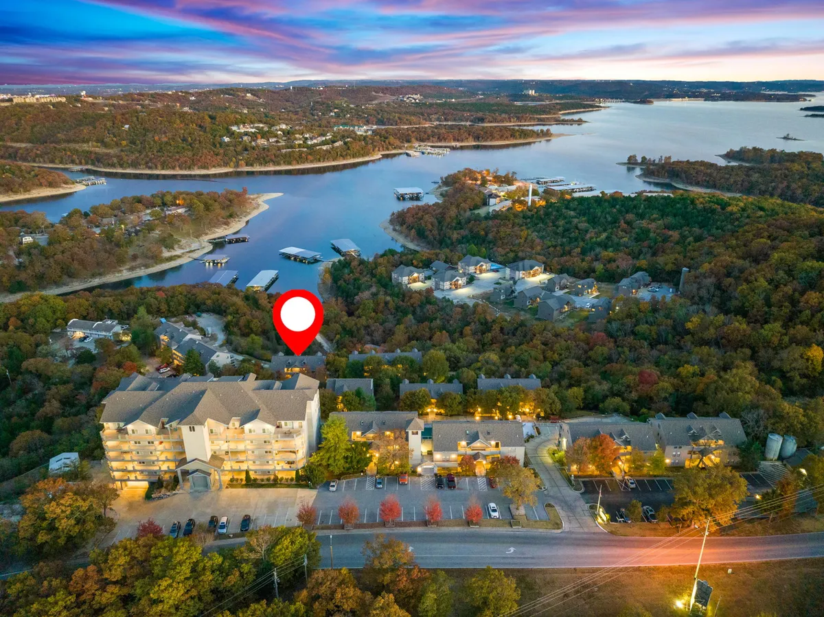 The image showcases a tranquil lakeside scene with calm water reflecting a stunning sunset. The foreground features a cluster of cozy condo buildings nestled among vibrant trees with autumn foliage, and boats dot the shoreline, inviting guests to enjoy the water. The atmosphere is peaceful, with hints of outdoor adventure and relaxation.