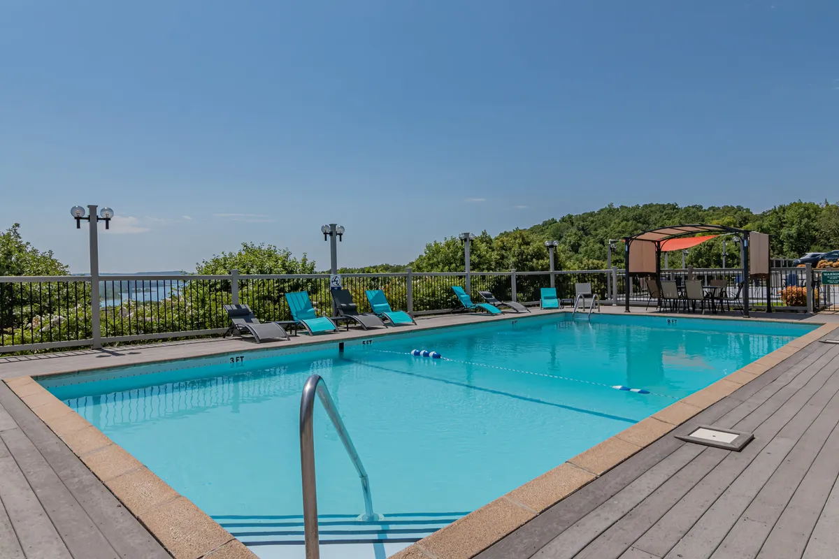 A sunny outdoor swimming pool area surrounded by lush greenery, featuring inviting lounge chairs, a canopy for shade, and space for groups to relax. The pool is central to the scene, making it an appealing focal point for vacation activities.