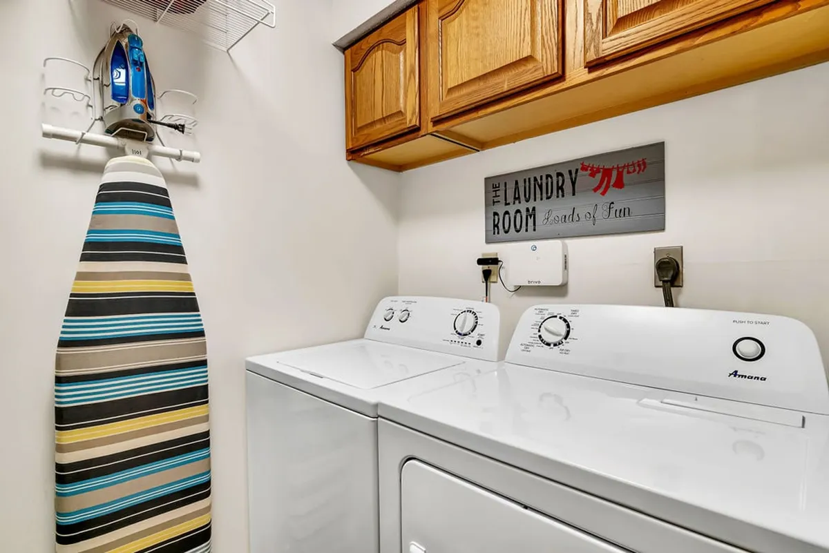 A practical laundry area with a washing machine, dryer, ironing board, and cheerful laundry room sign. The space is light-hearted and functional, making it easy to keep clothes fresh during your stay.