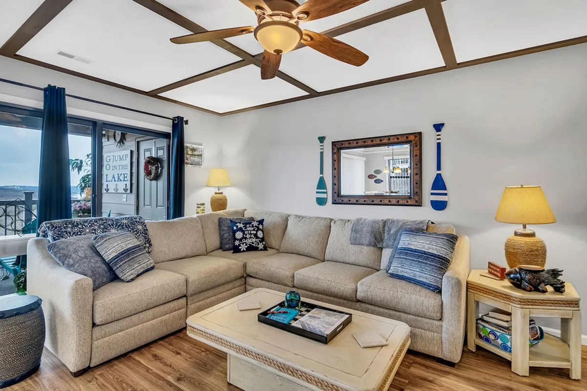 A close-up of the main seating area, featuring a sectional sofa, coffee table, and custom lake house decor. The room is bathed in soft lighting, fostering a cozy, comfortable atmosphere ideal for movie nights or board games. Blue accents and a decorative mirror add to the custom lake house feel, while the space feels inviting and perfect for gathering.