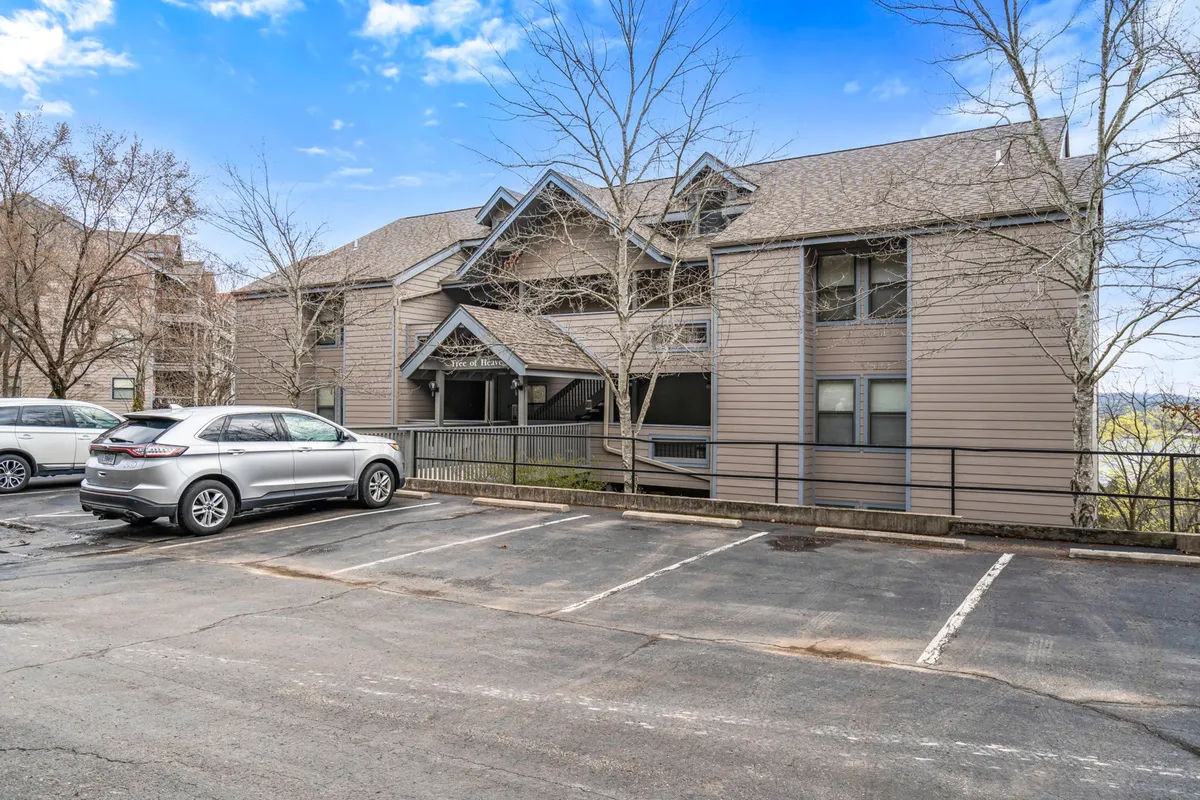 A wider view of the multi-level condo building, the spacious parking area, and leafy trees, giving a sense of the community setting and everyday convenience for guests. The setting feels accessible and friendly, ideal for families.