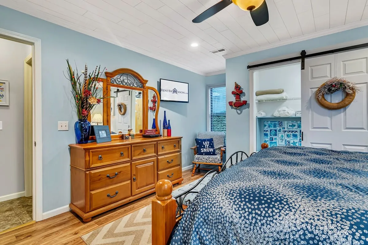 This interior photo features a cozy bedroom with a blue patterned bedspread, a wooden dresser topped with a mirror, nautical decorative accents, and a wall-mounted TV. Natural light streams in softly, creating a welcoming environment for relaxation or watching TV in privacy.