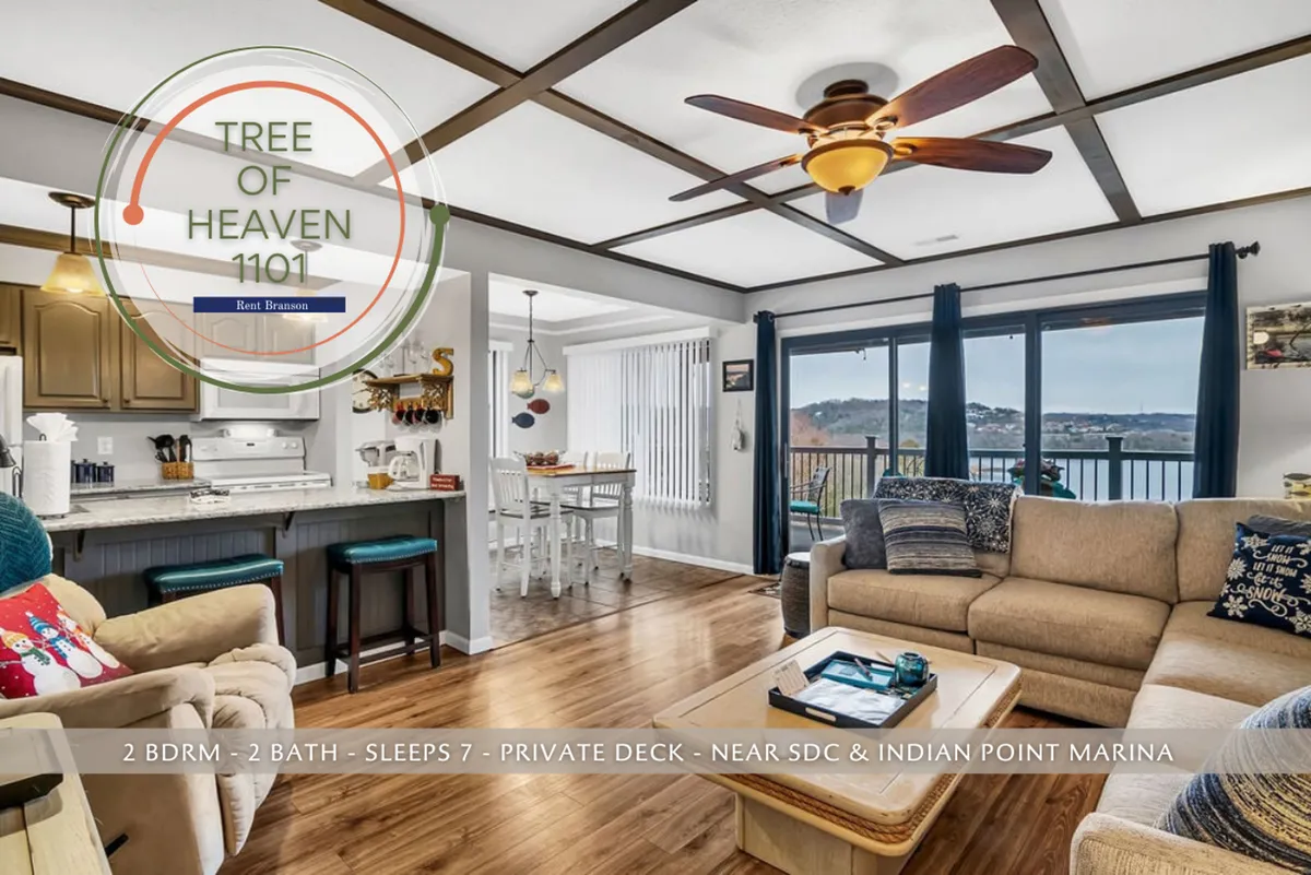 This image showcases an open-concept living space with a kitchen, living room seating, dining table, and large windows that offer an expansive view of the lake. The space is filled with natural light, and the arrangement encourages family gatherings and relaxation. The furnishings are plush and modern, and the overall ambiance is cozy and inviting, with direct visual connection to the outdoors.