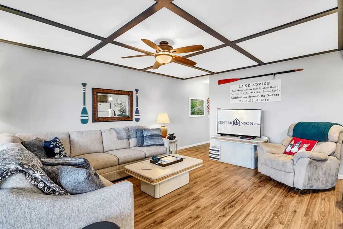 A cozy living room with a plush sofa, ceiling fan, decorative paddles, and a cheerful 'lake advice' sign. The space is bathed in soft light and offers an inviting setting for relaxation or socializing.