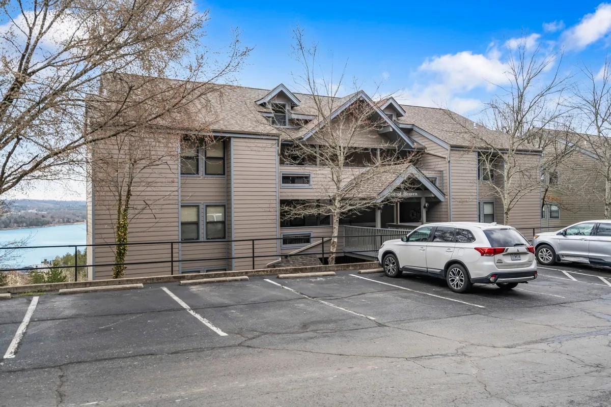 An exterior shot featuring the building facade, surrounded by trees, with a clear view of Table Rock Lake in the background. The parking lot is in the foreground, connecting the property to its natural environment and providing a sense of arrival.